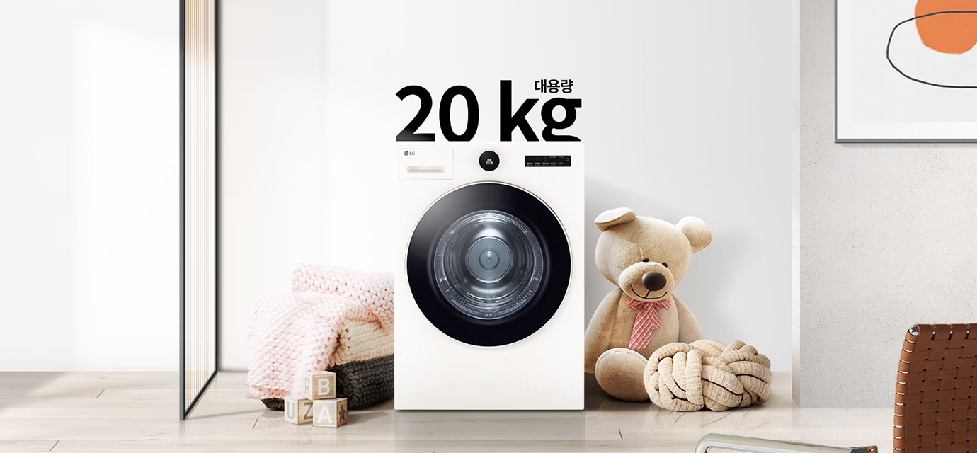 세탁기가 놓여 있으며, 상단에 대용량 20kg이라는 텍스트가 있습니다. 세탁기 주변에는 분홍색 니트 담요가 담긴 바구니와 곰 인형, 장난감 블록이 있습니다. 벽에는 추상적인 그림이 걸려 있습니다. 바닥은 나무 마루로 되어 있습니다.