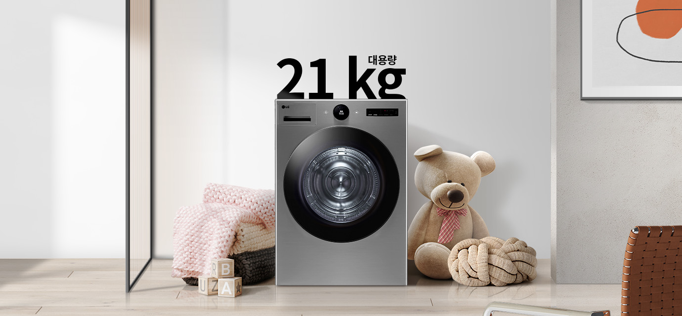 세탁기가 놓여 있으며, 21kg 대용량이라는 문구가 있습니다. 세탁기 주변에는 곰 인형과 두꺼운 니트 담요가 있고, 바닥에는 알파벳이 적힌 나무 블록이 있습니다. 벽에는 추상적인 그림이 걸려 있습니다.