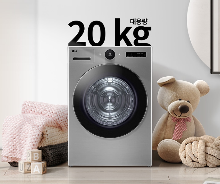 20kg 대용량 세탁기가 놓여 있으며, 주변에는 곰 인형과 장난감 블록, 그리고 핑크색 니트 담요가 보입니다. 세탁기 위에는 20kg 대용량이라는 텍스트가 있습니다. 배경은 깔끔한 실내 공간으로, 벽에는 추상적인 그림이 걸려 있습니다.