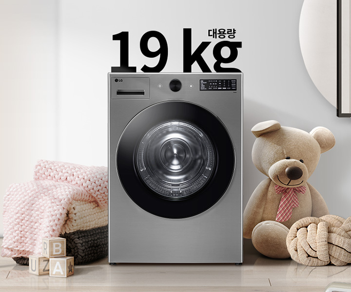 세탁기가 놓여 있으며, 상단에 대용량 19kg라는 글자가 있습니다. 세탁기 주변에는 곰 인형과 분홍색 담요가 놓여 있고, 바닥에는 B, U, A가 적힌 나무 블록이 있습니다. 배경은 밝은 색상의 벽과 그림이 걸려 있는 모습입니다.