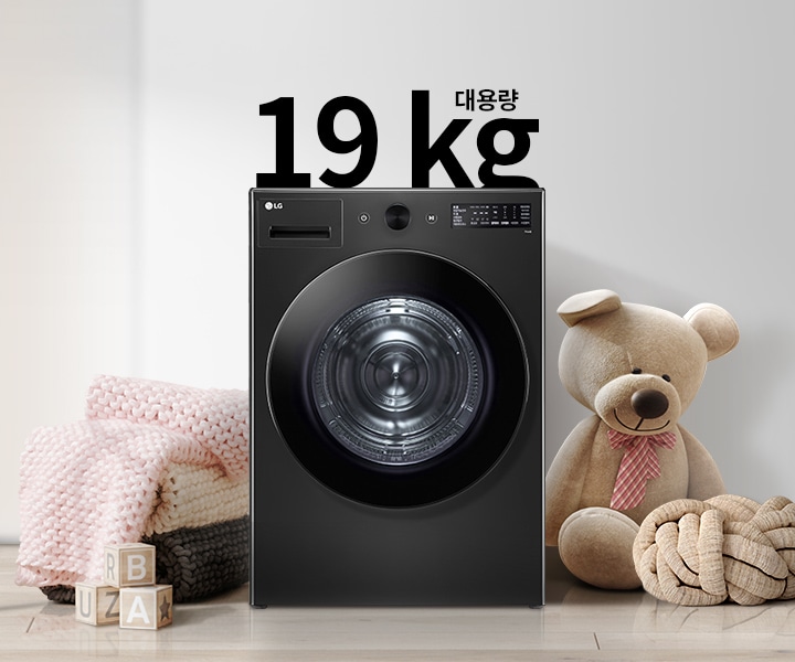 검정색 세탁기가 있으며, 그 위에 19kg 대용량이라는 텍스트가 있습니다. 세탁기 주변에는 분홍색 담요와 곰 인형이 놓여 있고, 나무 바닥 위에 알파벳 블록이 있습니다. 벽에는 추상적인 그림이 걸려 있습니다.