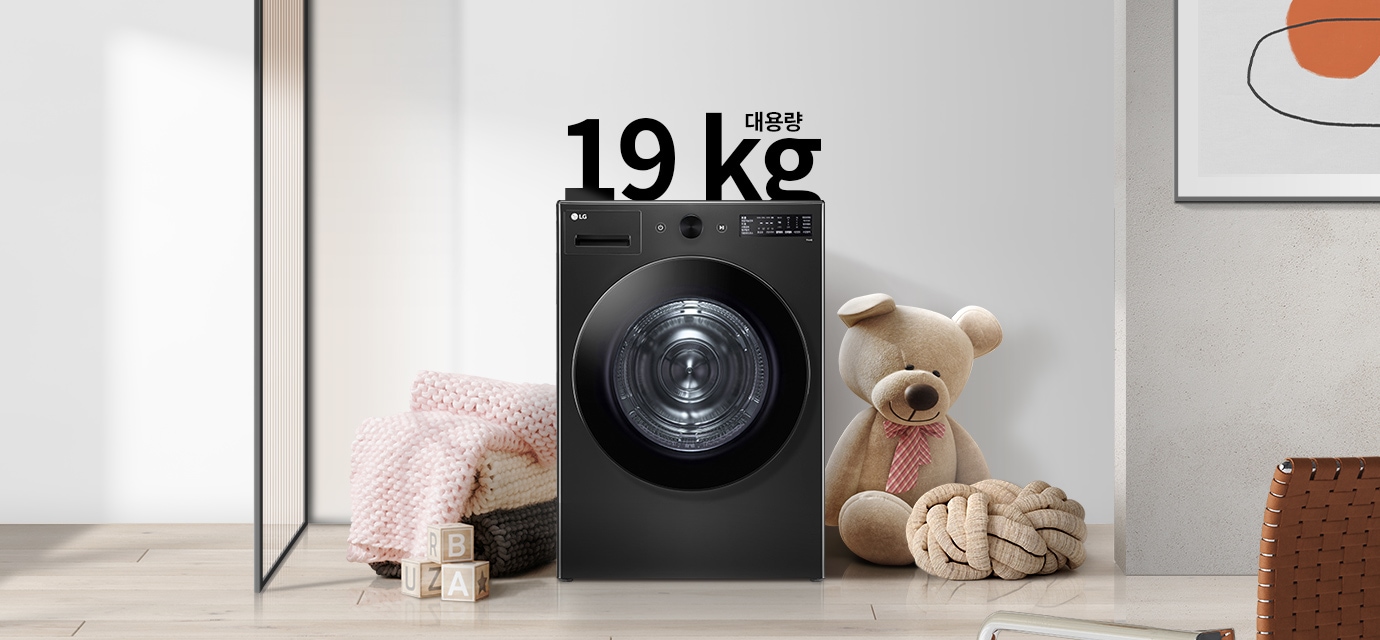 검정색 세탁기가 있으며, 그 위에 19kg 대용량이라는 텍스트가 있습니다. 세탁기 주변에는 분홍색 담요와 곰 인형이 놓여 있고, 나무 바닥 위에 알파벳 블록이 있습니다. 벽에는 추상적인 그림이 걸려 있습니다.
