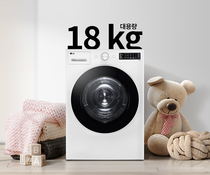 세탁기가 놓여 있으며, 18kg 대용량이라는 텍스트가 있습니다. 세탁기 옆에는 큰 곰 인형과 두꺼운 니트 담요가 놓여 있고, 바닥에는 알파벳 블록이 있습니다. 벽에는 추상적인 그림이 걸려 있습니다.