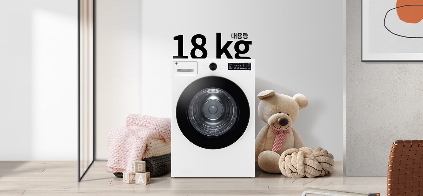 세탁기가 놓여 있으며, 18 kg 대용량이라는 문구가 있습니다. 세탁기 옆에는 곰 인형과 두꺼운 니트 담요가 놓여 있고, 바닥에는 알파벳 블록이 있습니다. 벽에는 추상적인 그림이 걸려 있습니다.