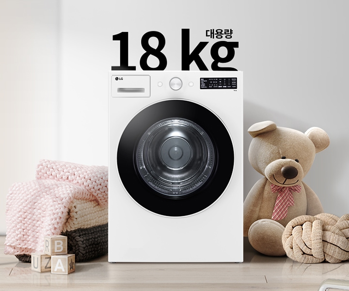 세탁기가 놓여 있으며, 18 kg 대용량이라는 문구가 있습니다. 세탁기 옆에는 곰 인형과 두꺼운 니트 담요가 놓여 있고, 바닥에는 알파벳 블록이 있습니다. 벽에는 추상적인 그림이 걸려 있습니다.