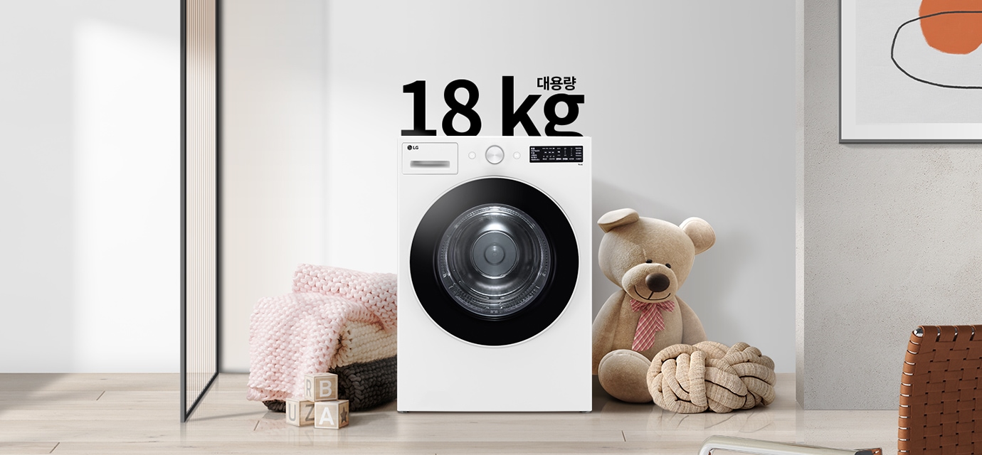 세탁기가 놓여 있으며, 18 kg 대용량이라는 문구가 있습니다. 세탁기 옆에는 곰 인형과 두꺼운 니트 담요가 놓여 있고, 바닥에는 알파벳 블록이 있습니다. 벽에는 추상적인 그림이 걸려 있습니다.