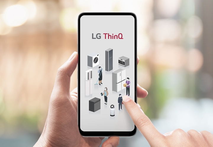 손에 들린 스마트폰 화면에 LG ThinQ라는 텍스트가 보이며, 다양한 가전제품과 사람들의 일러스트가 나타나 있습니다. 배경은 흐릿하게 처리되어 있습니다.
