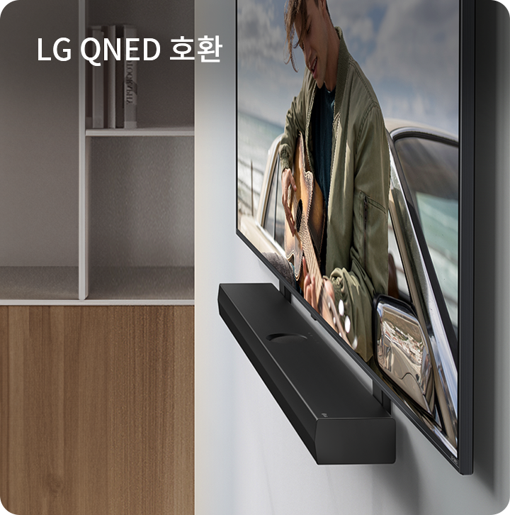 LG QNED 호환이라는 텍스트가 있으며, 벽에 설치된 TV와 사운드바가 보입니다. TV 화면에는 기타를 연주하는 사람이 있습니다. 배경에는 책장이 있습니다.