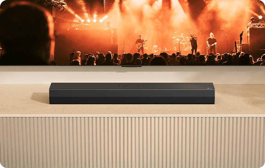 TV 앞 베이지색의 선반 위에 검은색 사운드바가 놓여져 있으며, TV 화면에서는 수많은 사람들이 강렬한 오렌지 빛 조명 아래 공연 중인 밴드를 바라보고 있는 모습이 보입니다.