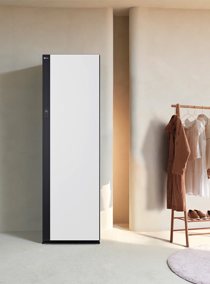 LG 로고가 있는 세련된 디자인의 흰색 가전제품이 놓여 있으며, 옆에는 다양한 색상의 옷이 걸린 나무 옷걸이가 있습니다. 바닥에는 신발과 바구니가 놓여 있고, 배경은 부드러운 색상의 벽과 파란색 의자가 배치되어 있습니다.