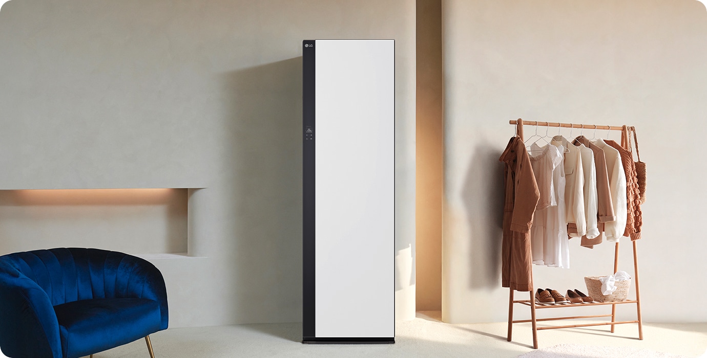 LG 로고가 있는 세련된 디자인의 흰색 가전제품이 놓여 있으며, 옆에는 다양한 색상의 옷이 걸린 나무 옷걸이가 있습니다. 바닥에는 신발과 바구니가 놓여 있고, 배경은 부드러운 색상의 벽과 파란색 의자가 배치되어 있습니다.