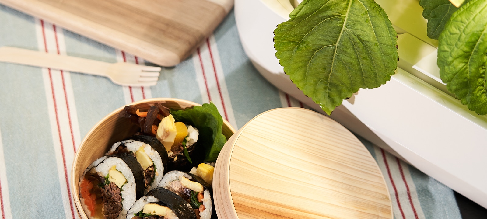 나무로 만든 도시락 통에 김밥이 담겨 있으며, 옆에는 나무 뚜껑이 놓여 있습니다. 김밥은 다양한 재료로 채워져 있고, 도시락 옆에는 커다란 녹색 잎이 보입니다. 배경에는 줄무늬가 있는 천과 나무 포크가 놓여 있습니다.
