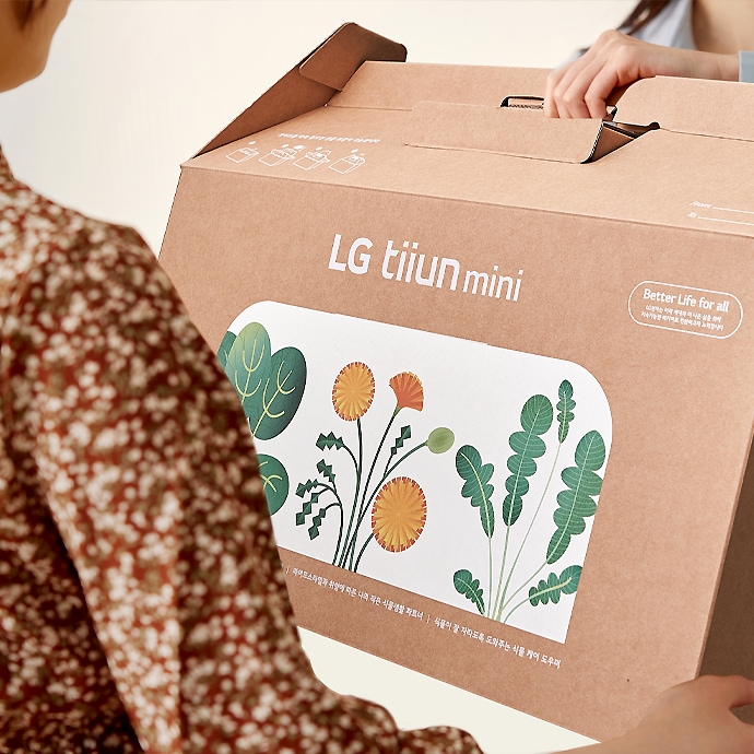 갈색_상자에_lg_tiiun_mini라는_글자가_인쇄되어_있으며,_상자에는_다양한_식물_그림이_그려져_있습니다._상자_옆에는_better_life_for_all이라는_문구가_적혀_있습니다._두_사람이_상자를_들고_있는_모습입니다.