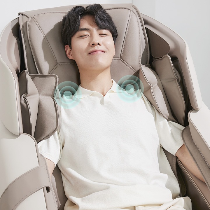 남성이 안마 의자에 앉아 눈을 감고 편안한 표정을 짓고 있습니다. 의자는 베이지색이며, 어깨 부분에 마사지 효과를 나타내는 파란색 그래픽이 있습니다. 남성은 흰색 셔츠를 입고 있습니다.