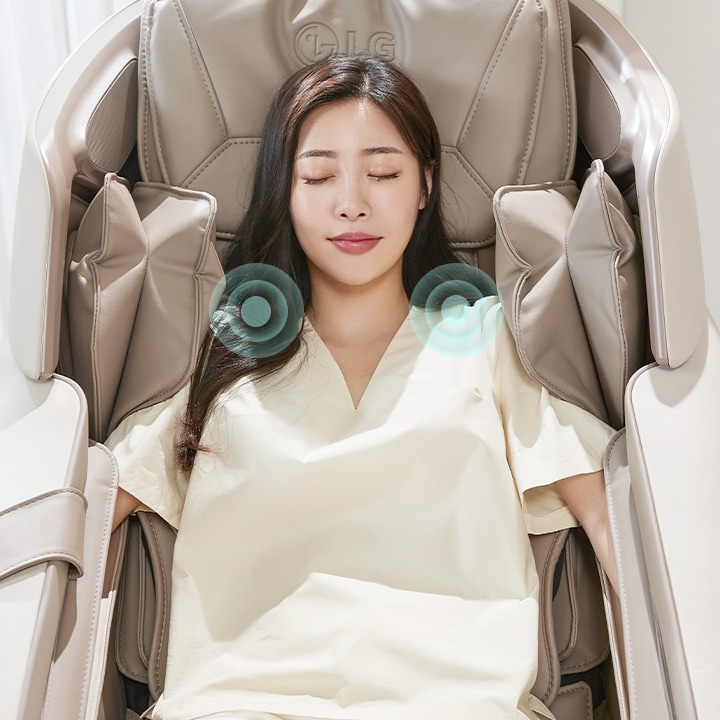 여성이 안마 의자에 앉아 눈을 감고 편안한 표정을 짓고 있습니다. 의자 상단에 'LG' 로고가 보이며, 어깨 부분에 마사지 효과를 나타내는 그래픽이 있습니다. 배경은 밝은 색상으로 되어 있습니다.