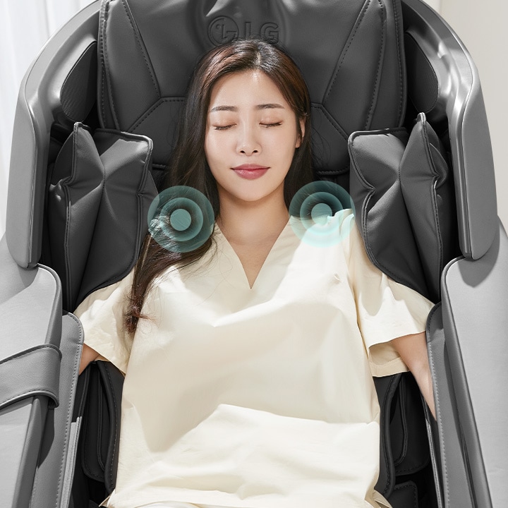 여성이_안마_의자에_앉아_눈을_감고_편안한_표정을_짓고_있습니다._어깨_부근에_소리나_진동을_나타내는_듯한_파란색_원형_그래픽이_보입니다._의자는_검은색이며,_주변은_밝은_색상의_실내입니다.
