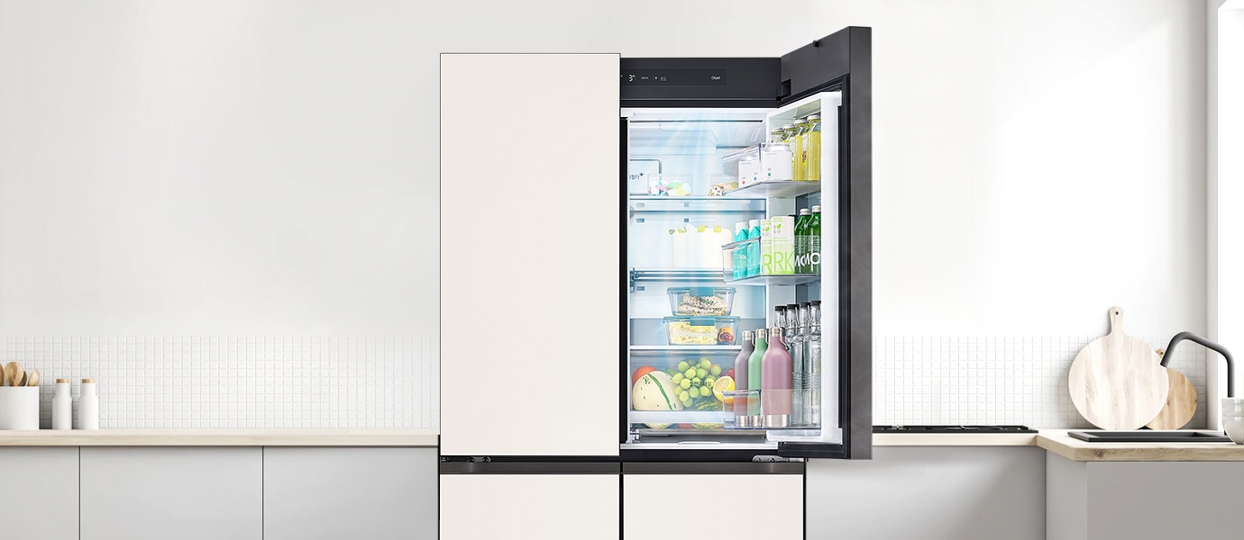 냉장고 문이 열려 있으며 내부에는 다양한 식품과 음료가 정리되어 있습니다. 선반에는 우유, 주스, 물병, 과일 등이 놓여 있고, 주방은 깔끔하게 정돈되어 있으며 흰색 타일 벽과 나무 조리대가 보입니다. 조리대 위에는 주방 도구와 병들이 놓여 있습니다.