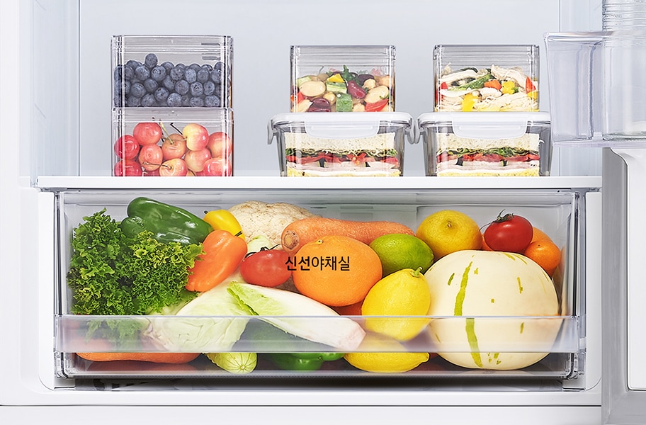 냉장고 내부에 다양한 과일과 채소가 정리되어 있습니다. 블루베리와 체리가 각각 투명한 용기에 담겨 있으며, 샌드위치와 채소가 층층이 쌓인 용기도 보입니다. 하단의 큰 서랍에는 상추, 피망, 당근, 오렌지, 레몬, 토마토, 멜론 등이 가득 차 있으며, 서랍에는 신선야채실이라는 글자가 적혀 있습니다.