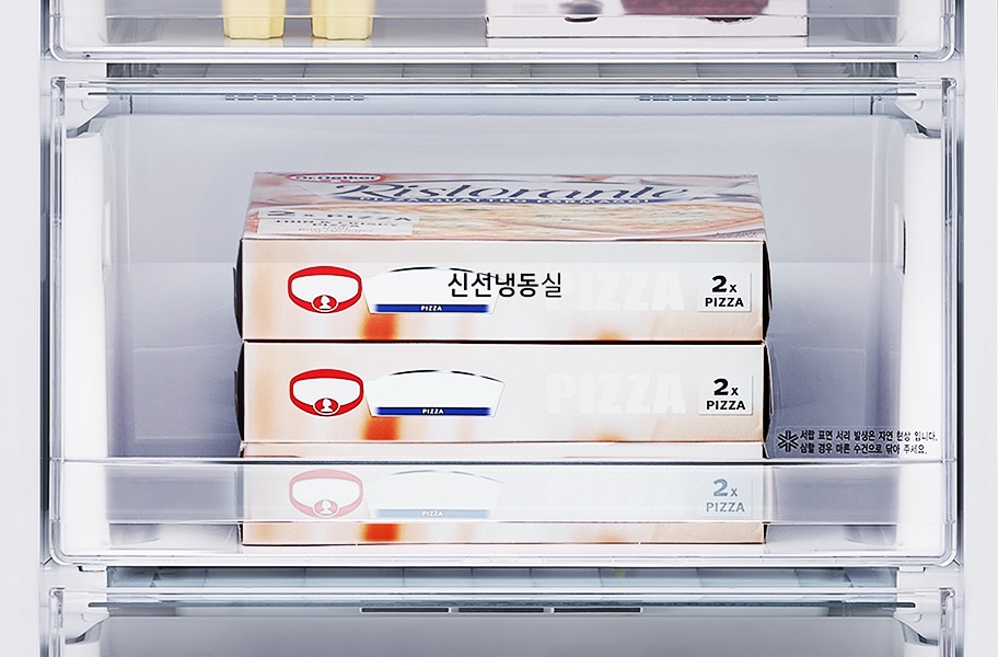 냉장고 안에 피자 상자 두 개가 쌓여 있습니다. 상자에는 신선냉동실과 2x PIZZA라는 글자가 보입니다. 상자 옆에는 색깔 코팅 시각 발생은 자연 현상입니다. 실물과 약간 다를 수 있으므로 유의 부탁이라는 문구가 적혀 있습니다. 상자 위쪽에는 다른 식품들이 보입니다.
