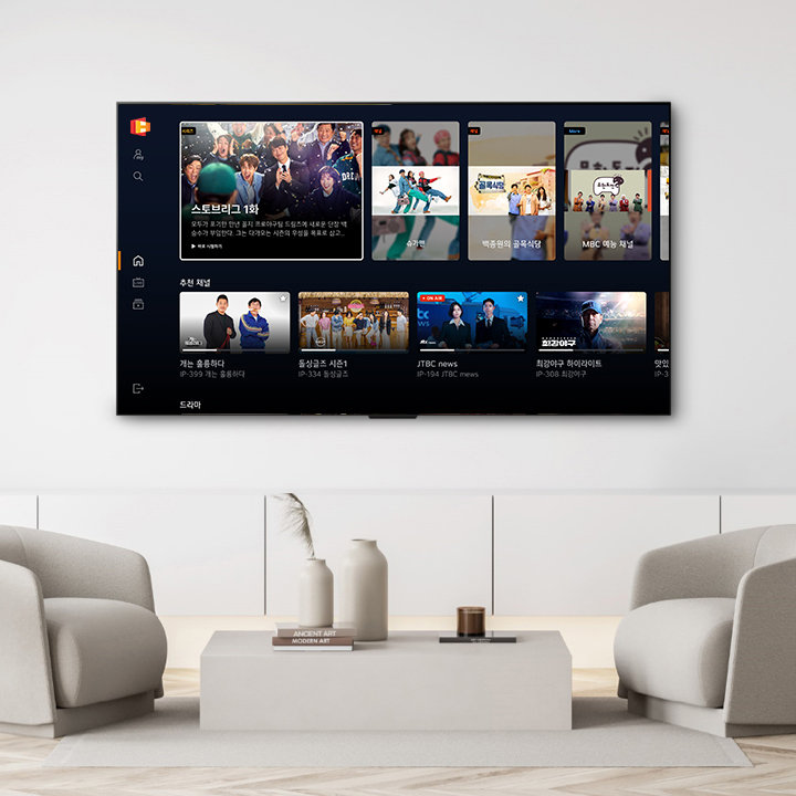 벽에 설치된 대형 TV 화면에 다양한 방송 프로그램 목록이 표시되어 있습니다. 화면에는 스토브리그 1화라는 프로그램이 강조되어 있으며, 그 외에도 가는 동안 한다, 동상이몽 시즌1, JTBC news, 최강의 하이라이트 등의 프로그램이 나열되어 있습니다. TV 아래에는 두 개의 소파와 중앙에 테이블이 놓여 있으며, 테이블 위에는 두 개의 화병과 책이 장식되어 있습니다.