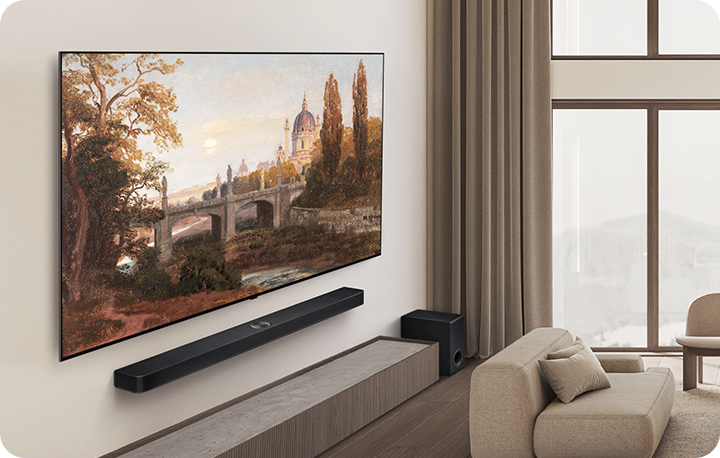 거실에 대형 TV가 벽에 걸려 있고, TV 화면에는 고풍스러운 풍경화가 표시되어 있습니다. TV 아래에는 사운드바가 설치되어 있으며, 그 옆에는 소파와 쿠션이 놓여 있습니다. 큰 창문을 통해 밝은 자연광이 들어오고 있습니다.