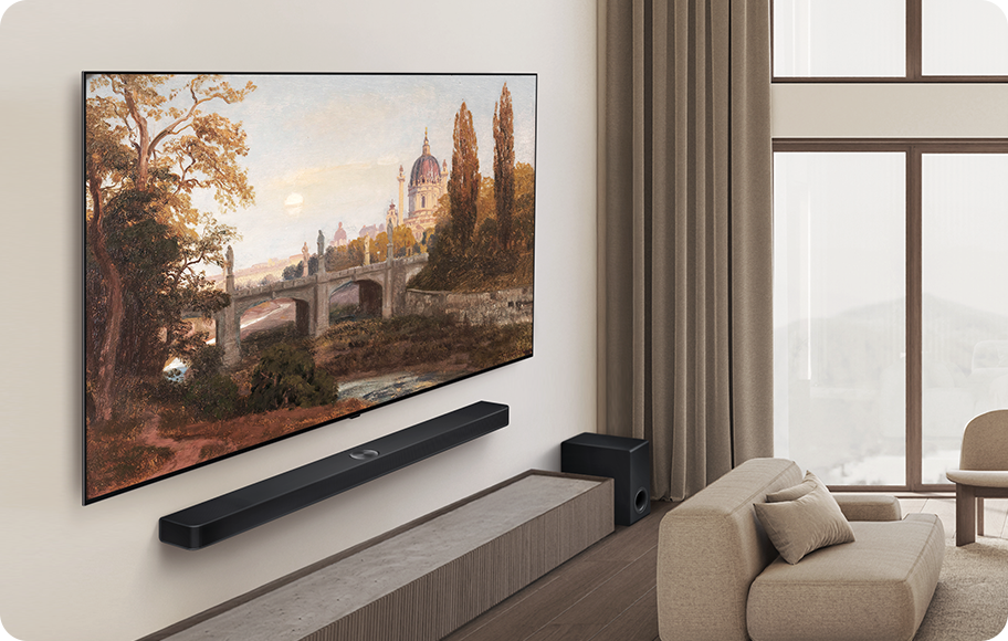 거실에 대형 TV가 벽에 걸려 있고, TV 화면에는 고풍스러운 풍경화가 표시되어 있습니다. TV 아래에는 사운드바가 설치되어 있으며, 그 옆에는 소파와 쿠션이 놓여 있습니다. 큰 창문을 통해 밝은 자연광이 들어오고 있습니다.