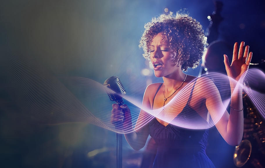 한 여성이 마이크를 잡고 노래를 부르고 있습니다. 그녀는 눈을 감고 있으며, 손을 들어 감정을 표현하고 있습니다. 배경은 흐릿하며, 음악의 파동을 나타내는 듯한 곡선형의 빛이 이미지에 더해져 있습니다. 조명은 부드럽고 따뜻한 분위기를 연출하고 있습니다.
