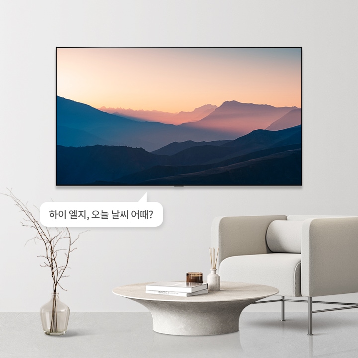 벽에 걸린 TV 화면에는 산의 실루엣이 보이는 일몰 장면이 담겨 있습니다. TV 아래에는 둥근 테이블과 소파가 놓여 있으며, 테이블 위에는 책과 장식품이 있습니다. 화면 밖 말풍선에는 하이 엘지, 오늘 날씨 어때?라는 텍스트가 포함되어 있습니다.