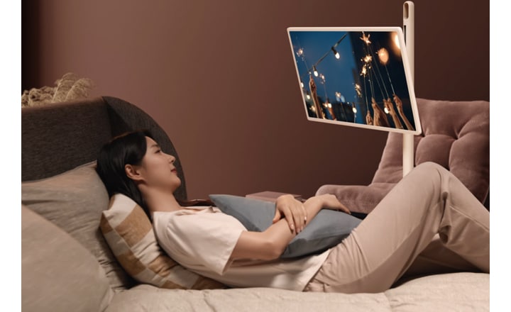 침대에 누워 있는 사람이 쿠션에 기대어 휴식을 취하고 있으며, 화면에는 밤하늘 아래 조명이 켜진 풍경이 보입니다. 주변은 따뜻한 색조의 조명으로 아늑한 분위기를 연출하고 있습니다.