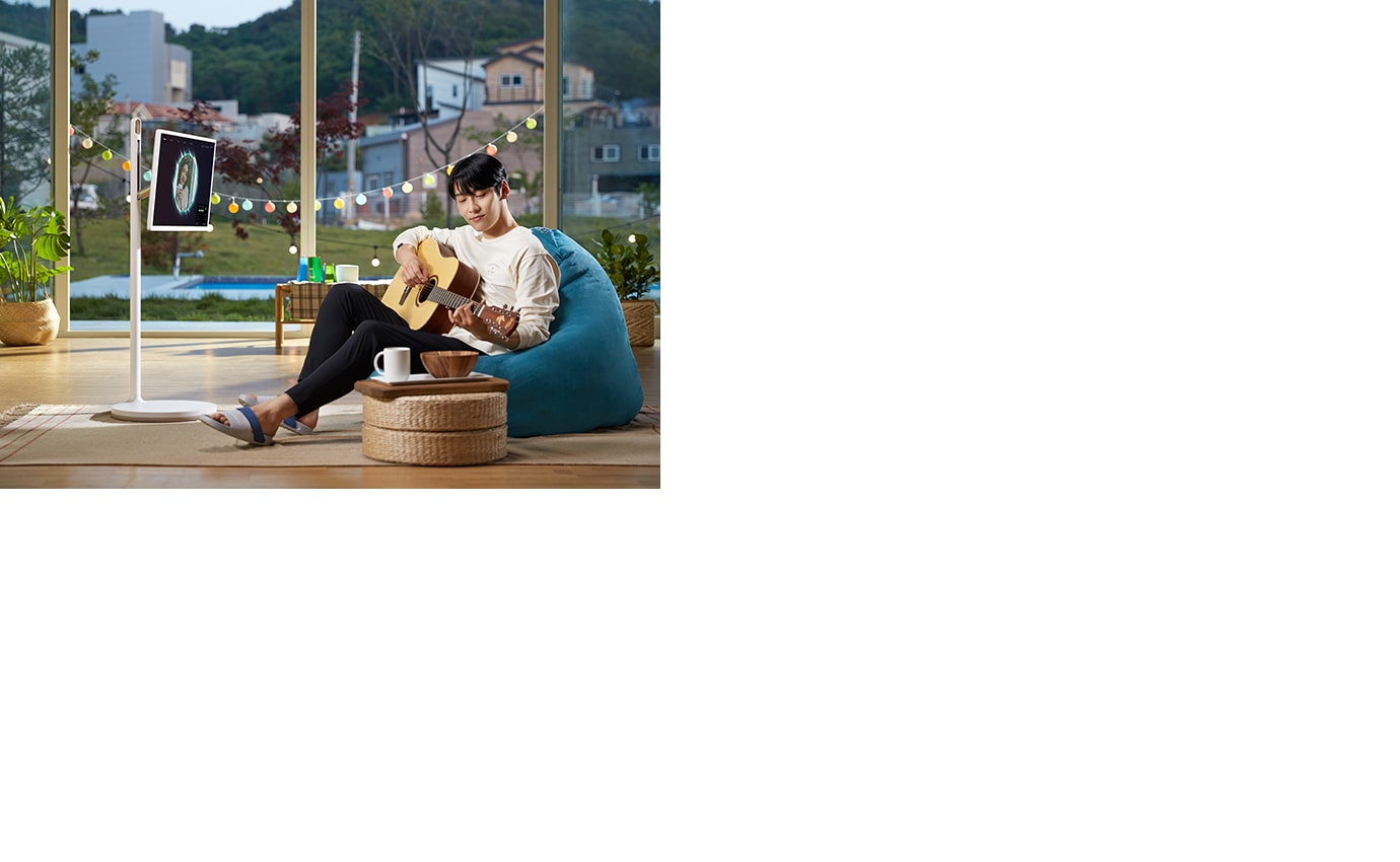 실내 공간에서 한 사람이 기타를 연주하고 있으며, 파란색 빈백에 앉아 있습니다. 주변에는 식물과 커피잔이 놓인 작은 테이블이 있습니다. 창밖으로는 주택과 나무가 보이며, 전구 장식이 걸려 있습니다. 스탠드에는 화면이 있으며, 화면에는 이미지가 표시되어 있습니다.