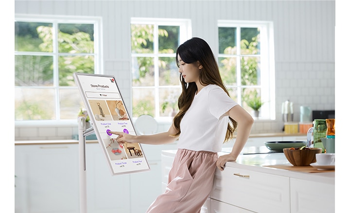 주방에서 한 여성이 디지털 스크린을 터치하고 있습니다. 스크린에는 Store Products라는 제목과 함께 여러 제품 이미지와 Product Info라는 텍스트가 보입니다. 배경은 밝고 창문을 통해 자연광이 들어오고 있습니다.