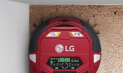 LG 로고가 있는 빨간색 로봇 청소기가 벽과 바닥이 만나는 곳에 위치해 있습니다. 청소기에는 디지털 디스플레이가 있으며, 다양한 아이콘과 숫자가 표시되어 있습니다. 청소기의 상단에는 주의 표시와 금지 아이콘이 있습니다. 바닥은 나무 질감입니다.