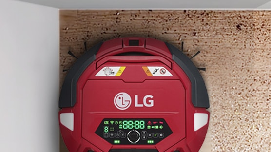 LG 로고가 있는 빨간색 로봇 청소기가 벽과 바닥이 만나는 곳에 위치해 있습니다. 청소기에는 디지털 디스플레이가 있으며, 다양한 아이콘과 숫자가 표시되어 있습니다. 청소기의 상단에는 주의 표시와 금지 아이콘이 있습니다. 바닥은 나무 질감입니다.