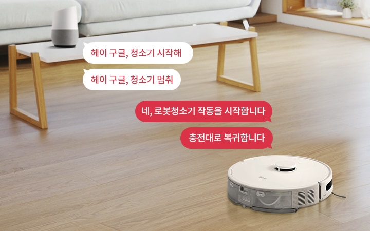 거실에 로봇청소기와 스마트 스피커가 있는 장면입니다. 스마트 스피커 옆에는 헤이 구글, 청소기 시작해와 헤이 구글, 청소기 멈춰라는 텍스트가 있습니다. 로봇청소기 옆에는 네, 로봇청소기 작동을 시작합니다와 충전대로 복귀합니다라는 텍스트가 있습니다. 바닥은 나무로 되어 있으며, 창가에는 식물이 놓여 있습니다.
