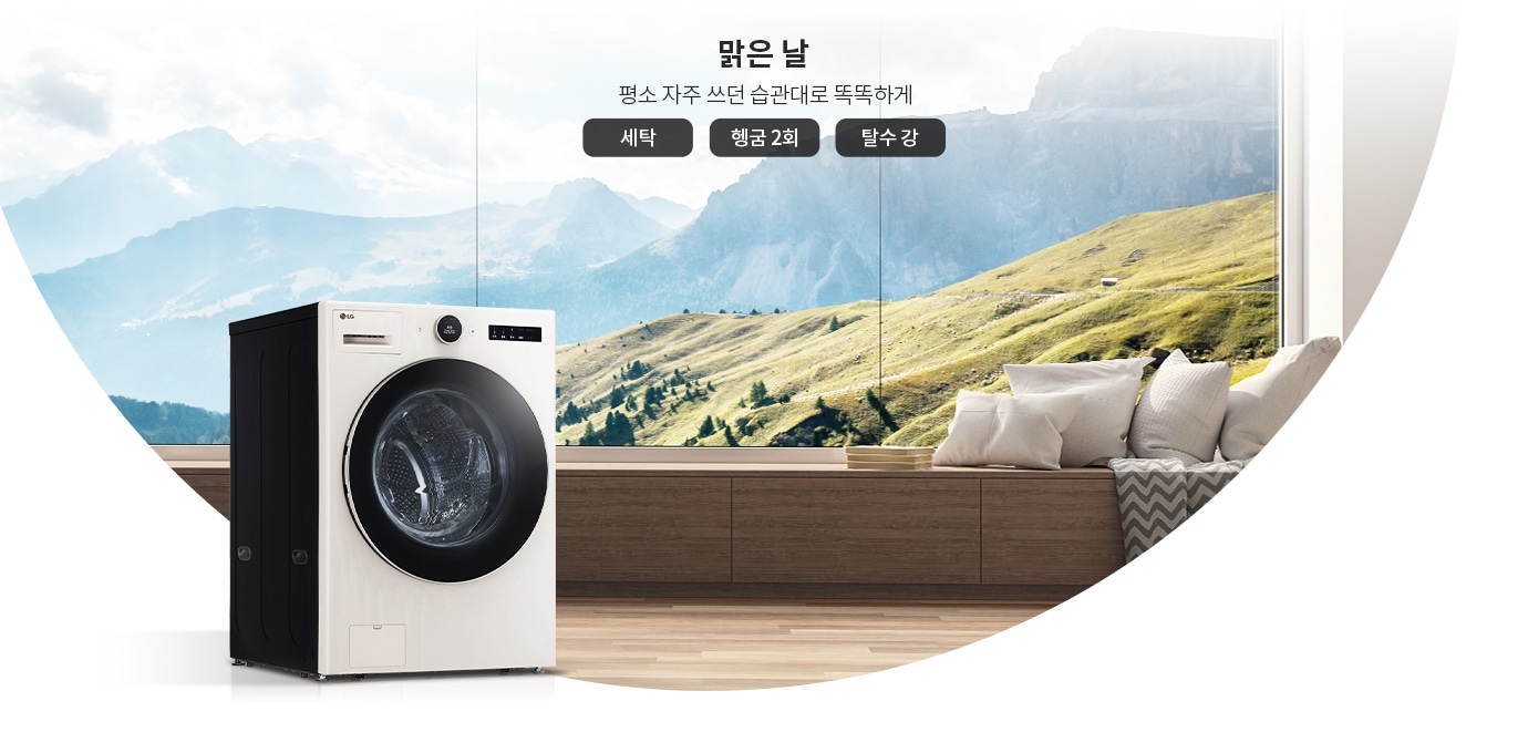 세탁기가 나무 바닥 위에 놓여 있으며, 뒤로는 큰 창문을 통해 산과 푸른 초원이 보입니다. 창문 옆에는 쿠션이 놓인 벤치가 있습니다. 