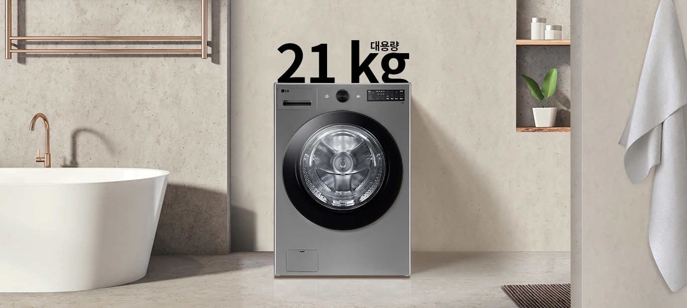 세탁기가 욕실에 놓여 있으며, 세탁기 위에는 21kg 대용량이라는 문구가 있습니다. 세탁기는 금속성 외관을 가지고 있고, 욕실에는 욕조와 수건이 걸려 있습니다. 벽에는 식물이 놓인 선반이 있습니다.