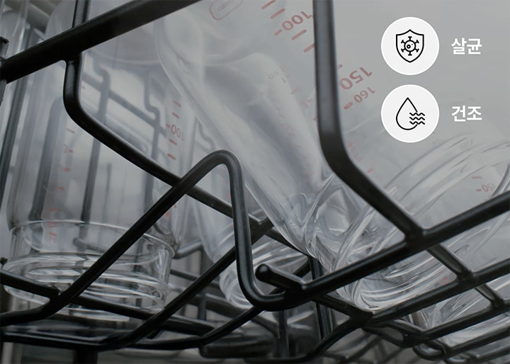 식기세척기 내부의 선반에 유리컵들이 거꾸로 놓여 있으며, 오른쪽 상단에는 두 개의 아이콘이 있습니다. 첫 번째 아이콘은 방패 모양 안에 살균을 상징하는 그림과 함께 살균이라는 글자가 있고, 두 번째 아이콘은 물방울 모양 안에 물결을 나타내는 그림과 건조라는 글자가 있습니다.