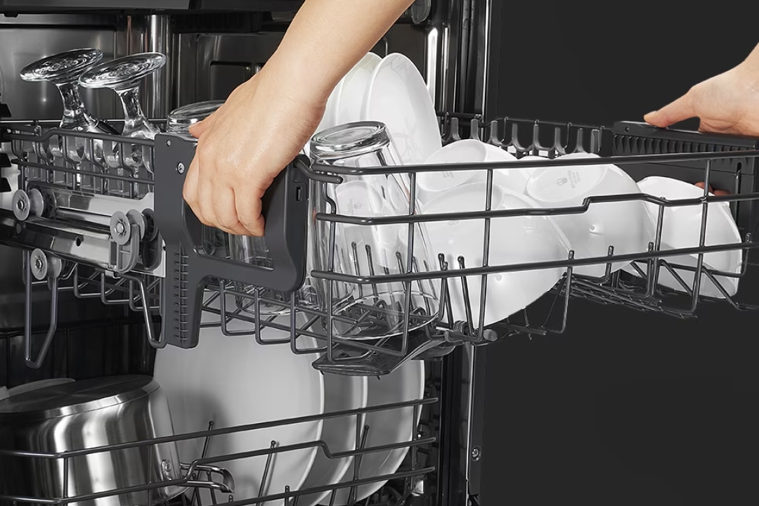 식기세척기 내부에서 손이 식기 선반을 당겨 내고 있으며, 위쪽 선반에는 흰색 컵이, 아래쪽 선반에는 흰색 접시가 놓여 있습니다.