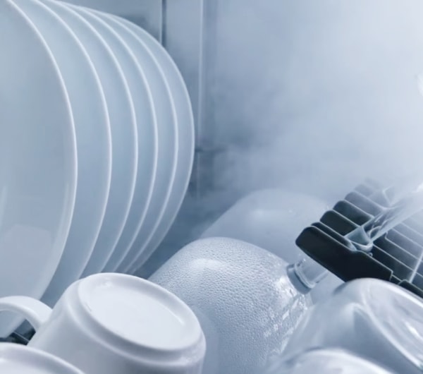 식기세척기 내부의 흰색 접시, 컵, 그리고 물방울이 맺힌 유리잔의 근접 사진으로, 수증기가 식으며 건조되는 모습입니다.