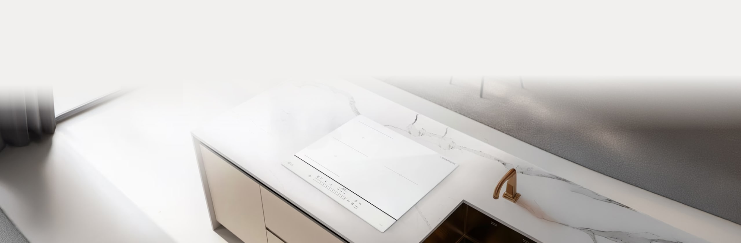 밝은 대리석 무늬의 조리대 위에 화이트 색상의 인덕션이 매립형으로 깔끔하게 설치되어 있습니다. 주변에는 싱크대와 골드 톤의 수전, 작은 그릇과 조미료 용기가 놓여 있어 밝고 모던한 주방 분위기를 완성합니다.