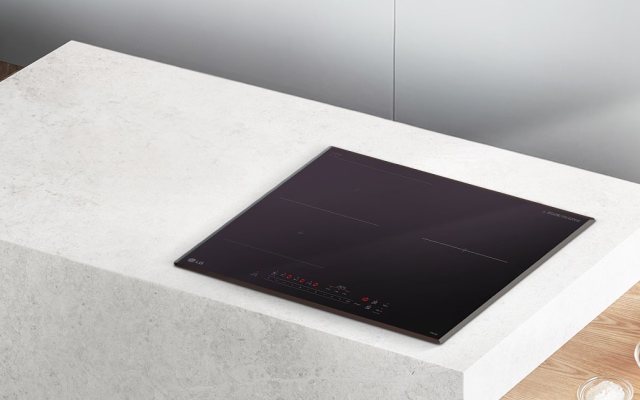 밝은 돌 질감의 조리대 위에 블랙 글라스 상판의 인덕션이 매립형으로 설치되어 있습니다. 주변에는 우드 바닥과 투명 볼에 담긴 달걀 등이 함께 보여 전체적으로 모던하고 따뜻한 주방 분위기를 연출합니다.