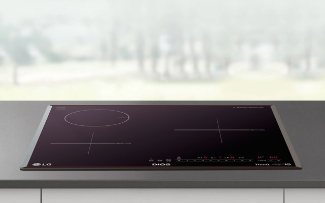 짙은 회색 조리대 위에 블랙 색상의 인덕션이 매립형으로 설치되어 있으며, 상판에는 원형·십자 형태의 조리 구역과 전원, 화력 조절, 타이머 등의 터치 아이콘이 붉은 색상으로 선명하게 표시되어 있습니다. 배경은 흐릿하게 보이는 밝은 외부 풍경으로 전체적으로 깔끔하고 모던한 주방 분위기를 연출합니다.
