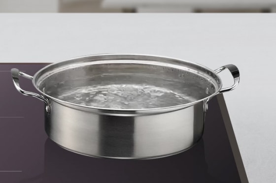 넓고 평평한 인덕션 조리대 위에 스테인리스 소재의 큰 냄비가 올려져 있으며, 냄비 안의 물이 끓어 거품과 잔물결이 생긴 모습입니다.