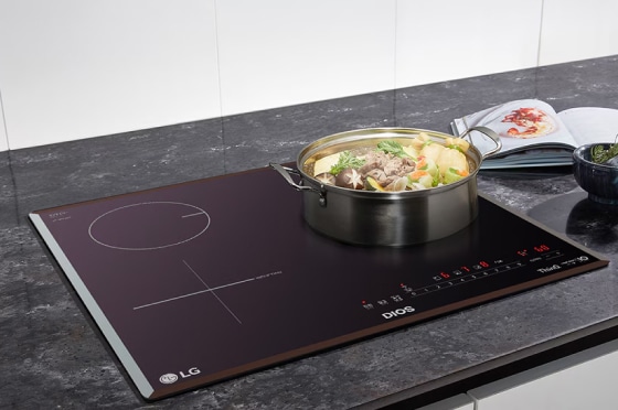 주방 조리대 위에 인덕션이 설치되어 있으며, 인덕션 위에는 다양한 채소와 고기가 들어 있는 냄비가 놓여 있습니다. 냄비 옆에는 요리책이 펼쳐져 있고, 작은 그릇과 반으로 잘린 레몬이 보입니다. 인덕션 표면에는 조작 패널과 함께 DIOS, ThinQ, Inverter 등의 텍스트가 있습니다. 배경은 대리석 느낌의 주방 상판입니다.