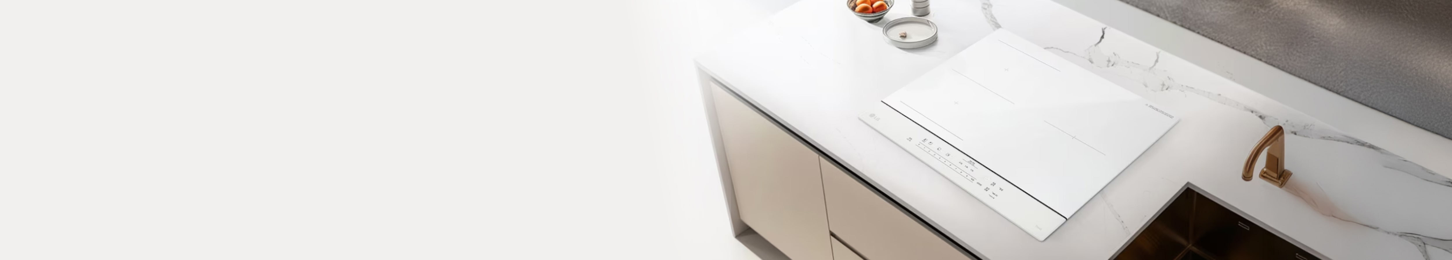 밝은 대리석 무늬의 조리대 위에 화이트 색상의 인덕션이 매립형으로 깔끔하게 설치되어 있습니다. 주변에는 싱크대와 골드 톤의 수전, 작은 그릇과 조미료 용기가 놓여 있어 밝고 모던한 주방 분위기를 완성합니다.