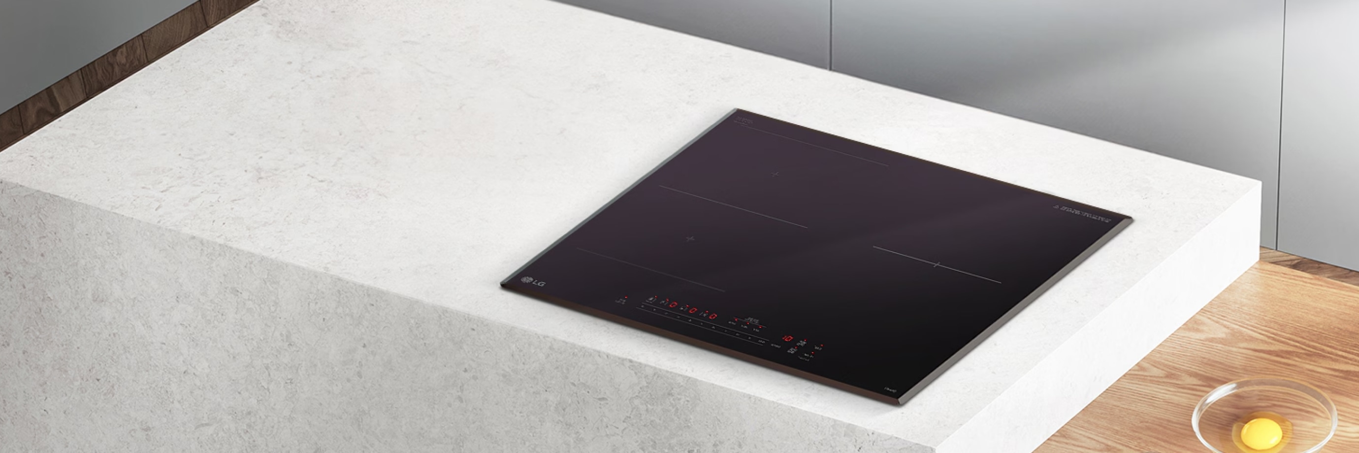 밝은 돌 질감의 조리대 위에 블랙 글라스 상판의 인덕션이 매립형으로 설치되어 있습니다. 주변에는 우드 바닥과 투명 볼에 담긴 달걀 등이 함께 보여 전체적으로 모던하고 따뜻한 주방 분위기를 연출합니다.