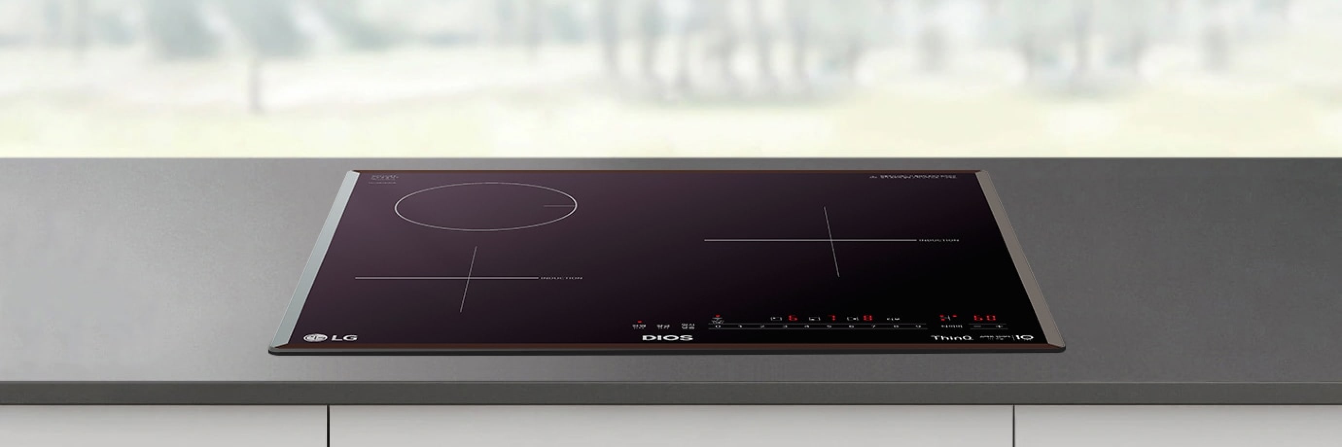 짙은 회색 조리대 위에 블랙 색상의 인덕션이 매립형으로 설치되어 있으며, 상판에는 원형·십자 형태의 조리 구역과 전원, 화력 조절, 타이머 등의 터치 아이콘이 붉은 색상으로 선명하게 표시되어 있습니다. 배경은 흐릿하게 보이는 밝은 외부 풍경으로 전체적으로 깔끔하고 모던한 주방 분위기를 연출합니다.