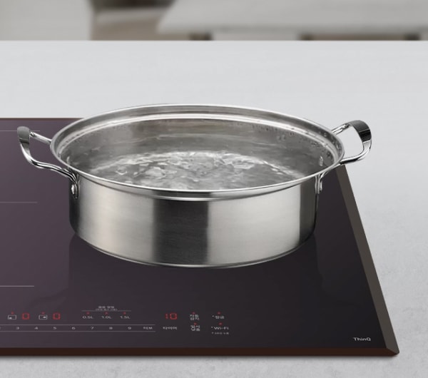 넓고 평평한 인덕션 조리대 위에 스테인리스 소재의 큰 냄비가 올려져 있으며, 냄비 안의 물이 끓어 거품과 잔물결이 생긴 모습입니다.