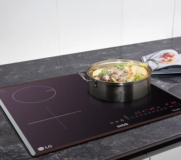 주방 조리대 위에 인덕션이 설치되어 있으며, 인덕션 위에는 다양한 채소와 고기가 들어 있는 냄비가 놓여 있습니다. 냄비 옆에는 요리책이 펼쳐져 있고, 작은 그릇과 반으로 잘린 레몬이 보입니다. 인덕션 표면에는 조작 패널과 함께 DIOS, ThinQ, Inverter 등의 텍스트가 있습니다. 배경은 대리석 느낌의 주방 상판입니다.