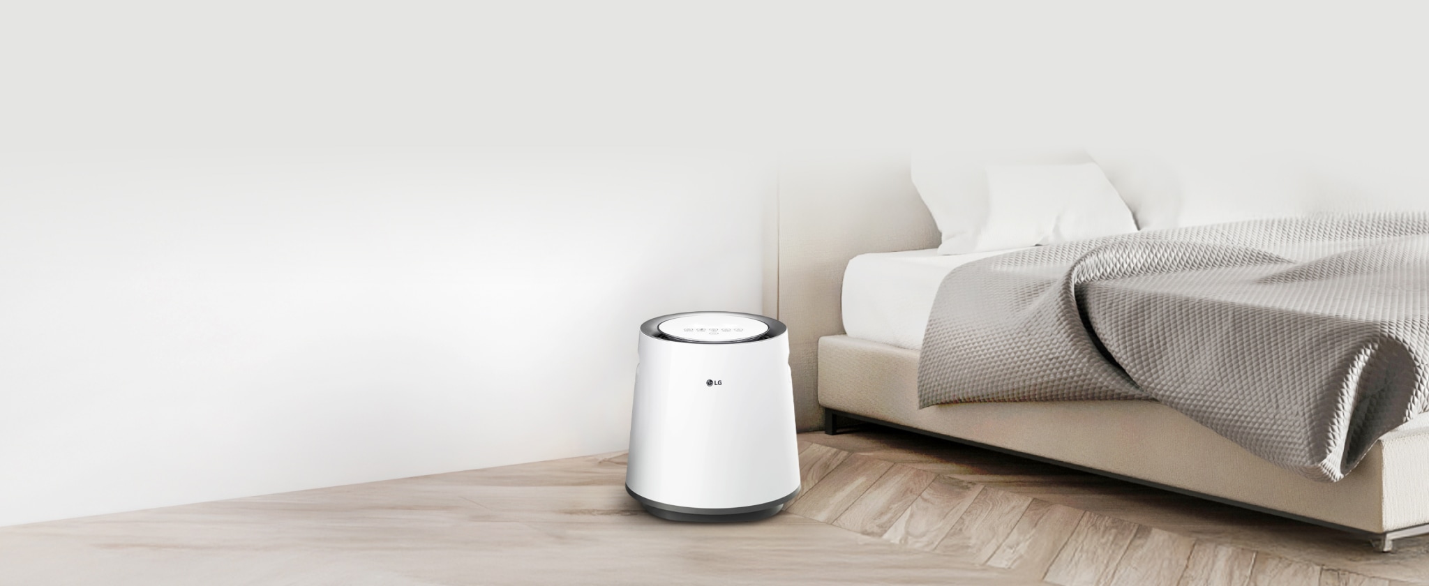 침실에 놓인 흰색 LG 퓨리케어 자연기화 가습기가 보입니다. 가습기는 원통형 디자인에 상단에 제어판이 있습니다. 침대는 베이지색 헤드보드와 흰색 침구가 있으며, 회색 담요가 놓여 있습니다. 방은 흰색 벽과 밝은 나무 바닥으로 꾸며져 있습니다.